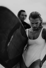 Load image into Gallery viewer, Euphoria Waves by Addison Jones Studio - a striking black-and-white photograph of a female surfer in a white swimsuit, showing emotions as she clutches her surfboard, captured at Wilmington Beach.