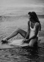 Load image into Gallery viewer, A stunning black and white photograph titled 'Riding Tide,' showcasing a woman in a white swimsuit sitting on a surfboard at Wilmington Beach, North Carolina, capturing the essence of serene ocean vibes.
